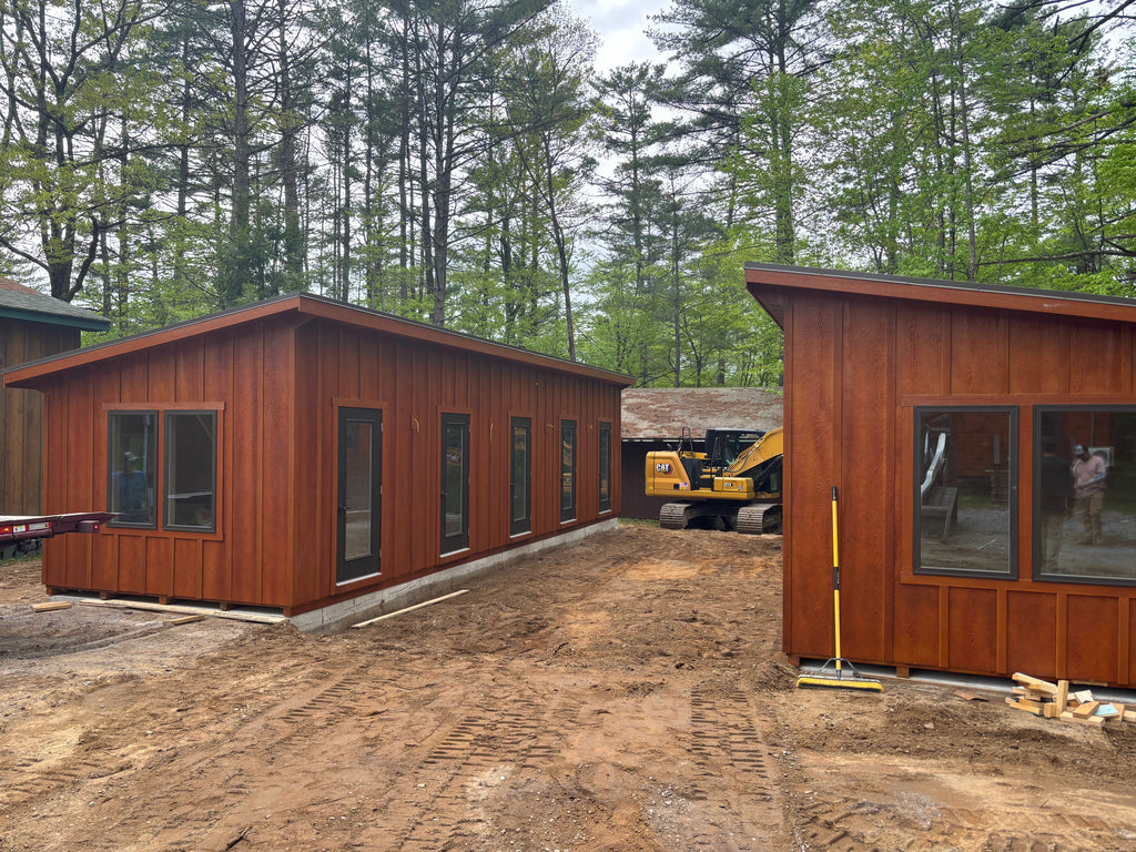 Two wooden modular buildings on a construction site with trees in the background.